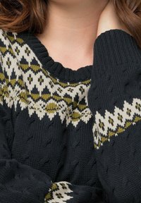 Stickad tröja med en mörk marinblå bas och ett strukturerat mönster, vita och olivgröna geometriska mönster över bröstet och ärmarna.