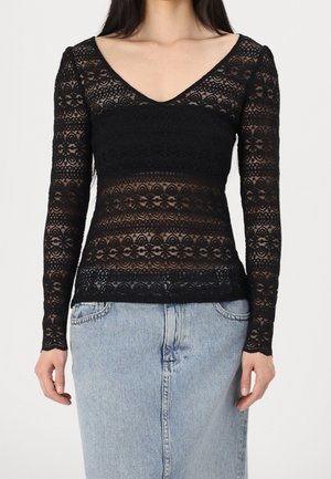Kvinde iført en tætsiddende sort blondebluse med lange ærmer, blomsterprint og V-udskæring, kombineret med en lyseblå denim nederdel.