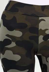 Högmidjade leggings i ett mjukt, elastiskt tyg som har ett mörkgrönt och svart kamouflage-mönster med en slät, matt yta.