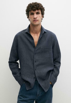 Uomo con capelli ricci che indossa una camicia grigio scuro con bottoni sopra jeans blu, in piedi con le mani nelle tasche su uno sfondo bianco semplice.