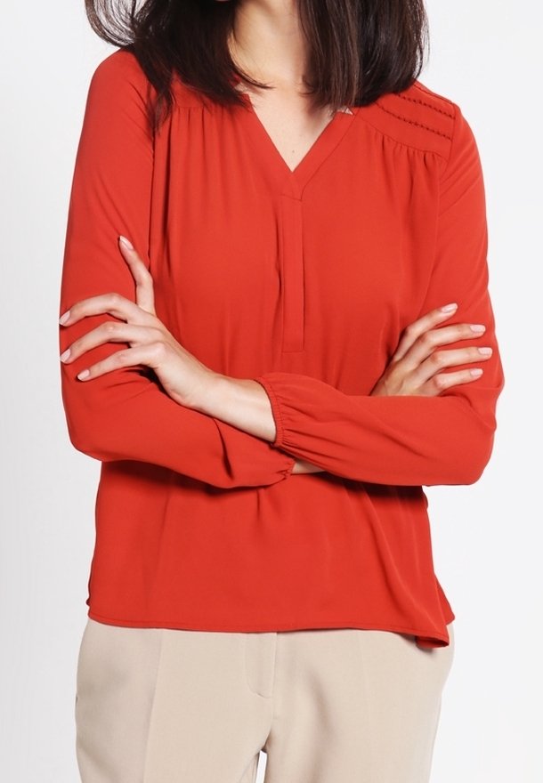 Blouse rouge avec un design en V, manches longues avec des poignets élastiques et des plis subtils aux épaules. Texture du tissu lisse.