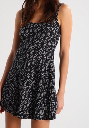 Femme portant une robe noire sans manches avec des motifs géométriques blancs, cintrée à la taille et évasée au niveau de la jupe, sur un fond uni.