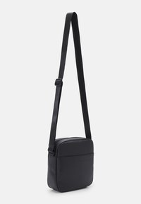 Crni crossbody torba od glatkog materijala, s rektangularnim oblikom, zatvaranjem na zip i podesivim remenom. Kompaktni dizajn s prednjim džepom.