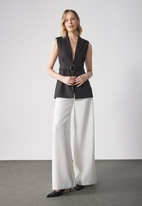 Schwarze ärmellose Blazerjacke mit tiefem V-Ausschnitt und goldener runder Gürtelschnalle, kombiniert mit weißen weit geschnittenen Hosen und schwarzen High Heels.