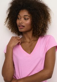 Roze V-hals T-shirt met korte mouwen, gemaakt van een lichtgewicht, zachte stof; model heeft krullend haar en een ontspannen houding tegen een witte achtergrond.