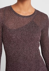 Gros plan d'une personne portant un haut violet transparent à manches longues côtelé par-dessus un soutien-gorge assorti.