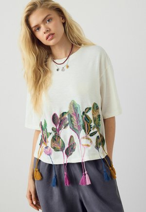 Tânără cu păr blond lung poartă un top alb cu mâneci scurte, cu frunze brodate colorate și ciucuri, asortat cu pantaloni închiși la culoare.