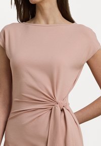 Top casual rosa con maniche corte e dettaglio annodato davanti in vita, realizzato in tessuto morbido e elasticizzato con una texture liscia.