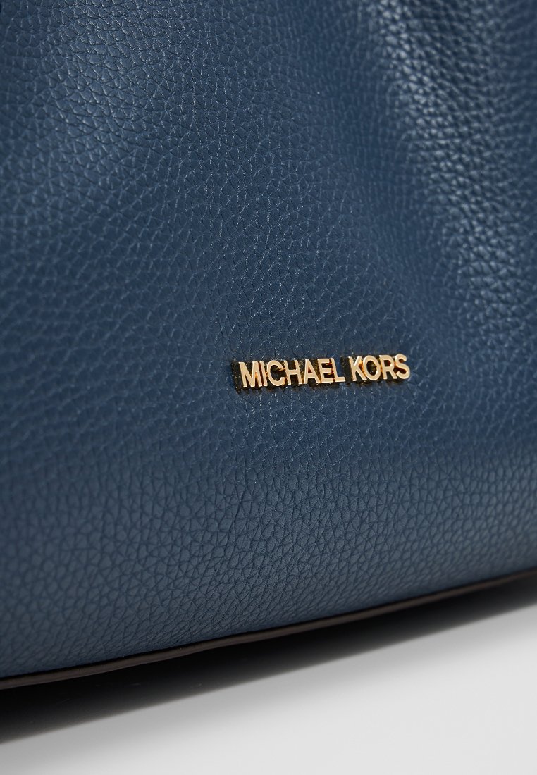 Sac à main en cuir bleu en relief avec une plaque logo "MICHAEL KORS" en ton doré. La texture est gaufrée, offrant une surface tactile.