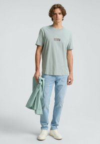 Jasnozielona bawełniana koszulka z małym nadrukiem na piersi, w połączeniu z jasnoniebieskimi dżinsami i białymi trampkami, trzymając pasującą zieloną koszulę.