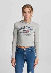 Felpa grigia corta con cappuccio foderato e orlo ondulato, con la scritta 'NEW YORK' e una stampa della bandiera americana. Indossata con jeans blu.