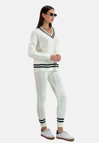 Jersey de punto grueso blanco y pantalones a juego con detalles de franjas negras en los puños y el dobladillo. La modelo lleva gafas de sol negras elegantes y zapatillas blancas.