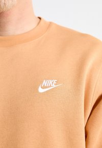 Närbild av en beige sweatshirt med vit Nike-logotyp broderad på vänster bröst, bärs av en person med ljus hy och blond skägg.