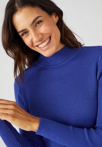 Pullover a collo alto blu a coste con maniche lunghe, caratterizzato da un design aderente e un tessuto testurizzato. Inquadratura ravvicinata di una persona che sorride.