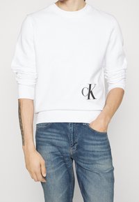 B ílá mikina se zaobleným výstřihem, ribbovanými manžetami a lemem. Na dolní levé straně se nachází malé černé logo "ck". Nosí se s modrými džíny.