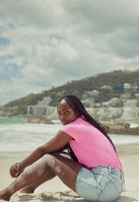 Model nosi svijetloružičastu majicu kratkih rukava i svijetloplave traper kratke hlače, sjedi na pješčanoj plaži s valovima u pozadini.