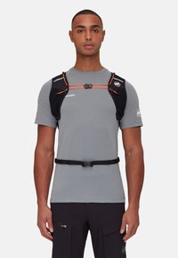 Chaleco de running negro con detalles naranjas, correas ajustables y un broche. Lleva encima una camiseta gris, con bolsillos para almacenamiento.