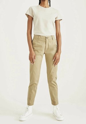 Vrouw staand met een effen beige T-shirt, tan hoge taille broek en witte high-top sneakers tegen een lichte achtergrond.