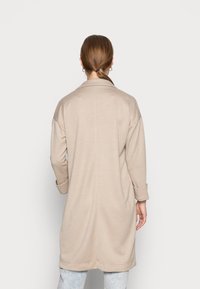 Woman with brown hair in a low ponytail wearing a beige long coat and light blue jeans, facing away against a plain light background.