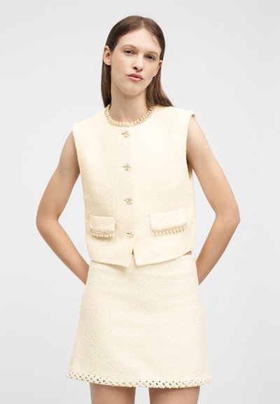 Γυναίκα που φοράει αμάνικο γιλέκο από κρεμ τουίντ με πέρλες κουμπιά και ταιριαστό μίνι φούστα με διακοσμητική επένδυση, στέκεται με τα χέρια στη μέση.