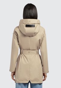 Beige jacket with a hood, cinched waist, and a belted design. Features a logo patch on the back and a smooth, water-resistant material.