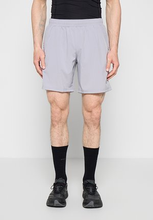 Homme portant un short de sport gris clair, des chaussettes noires et des chaussures de course noires, debout devant un fond gris clair uni.