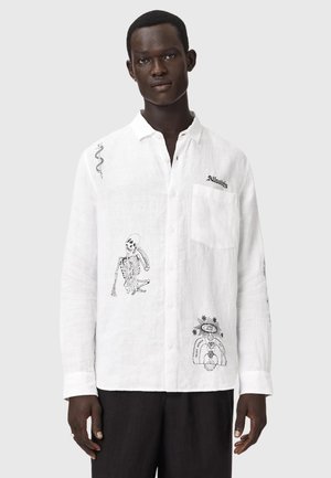 Mężczyzna w białej koszuli na guziki z czarnymi ilustracjami szkieletu i motywami niebiańskimi, w czarnych spodniach, stojący na tle jednolitego tła.