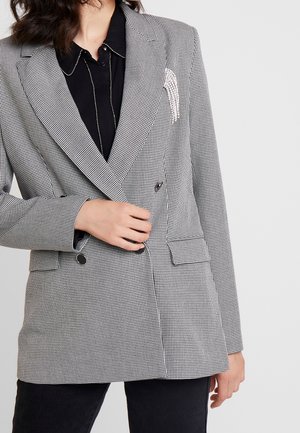 Nainen pukeutunut mustavalkoiseen kaksoisrintaisen piikkikuviolliseen bleiseriin, jossa hopeinen koriste liepeessä, sekä mustaan napitettuun paitaan.