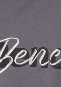 Grijze textiel met "Benefit" in grote, zwarte en textuur-witte borduring. Het lettertype heeft een soepele, vloeiende uitstraling.