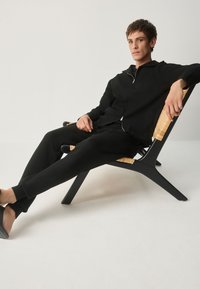 Jachetă neagră cu fermoar și pantaloni din material moale, așezată pe un scaun negru din lemn cu accente împletite. Modelul adoptă o postură casual.