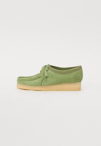 Chaussure en daim verte avec un bout arrondi, des détails cousus, un lacet noué à l'avant et une semelle en caoutchouc beige.