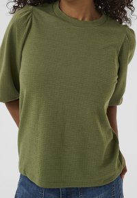 Maglietta verde oliva a maniche corte con un tessuto texturizzato, caratterizzata da un collo rotondo e una leggera arricciatura sulle spalle. Abbinata a denim.