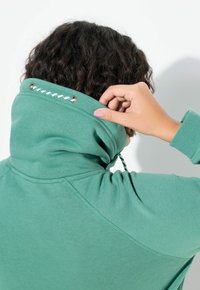 Tyrkysová mikina s vysokým límcem, vybavená stahovací šňůrkou protaženou zlatými očky. Látka je hladká s jemnou texturou.