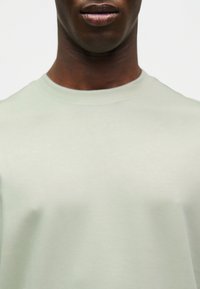 Κοντινό πλάνο του κάτω μέρους του προσώπου και του λαιμού ενός ατόμου που φοράει ανοιχτόχρωμο μπλουζάκι με στρογγυλή λαιμόκοψη σε απλό φόντο.