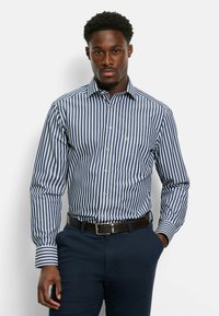Camicia a righe blu e bianche con colletto, maniche lunghe e tasca sul petto. Indossata con pantaloni scuri e una cintura marrone.