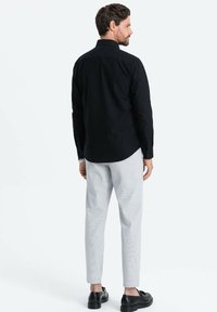 Camicia nera a maniche lunghe con colletto, abbinata a pantaloni grigi chiari con motivo e scarpe nere, vista da dietro.