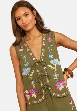 Kvinde iført olivengrøn ærmeløs vest med farverig blomsterbroderi og bindebånd, accessoriseret med store armbånd.