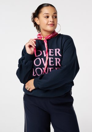 Pull bleu marine avec des manches bouffantes surdimensionnées, arborant un texte rose en gras qui dit "OVER LOVE". Le mannequin porte un foulard rose à motifs autour du cou.