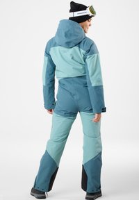 Ski suit in teal tones featuring a hood, contrasting panels, and waterproof material. Adjustable cuffs and zippered pockets are visible.