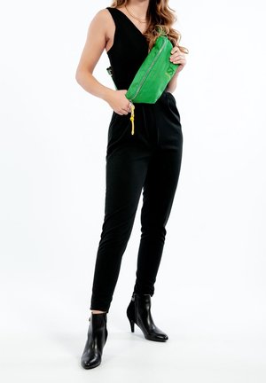 Tuta nera con gambe aderenti, abbinata a una cintura a tracolla verde con texture, dotata di zip argentata e dettagli in hardware giallo. Stivali neri alla caviglia.