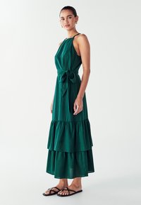Zelena, višeslojna maxi haljina s vezanjem u struku, izrađena od lagane tkanine. Ima tanke naramenice i lepršavi kroj. Uparena s crnim sandalama.