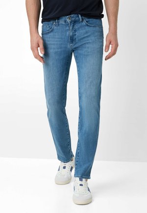 Mann trägt mittelblaue Slim-Fit-Jeans und weiße Turnschuhe mit marineblauen Streifen, steht vor weißem Hintergrund.