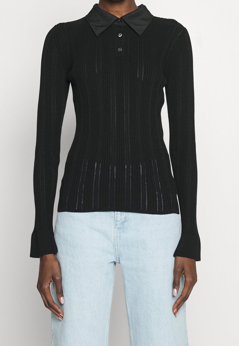 Femme portant un haut noir côtelé à manches longues ajusté avec un large col et trois boutons, associé à un jean taille haute bleu clair.