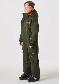 Grön vattentät skidoverall med orange detaljer, fickor med dragkedja och hög krage. Materialet verkar tåligt; lämpligt för kalla väderaktiviteter.