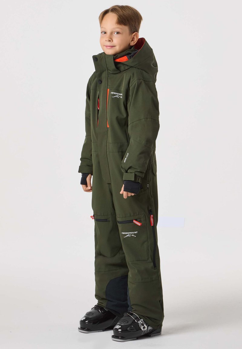 Grön vattentät skidoverall med orange detaljer, fickor med dragkedja och hög krage. Materialet verkar tåligt; lämpligt för kalla väderaktiviteter.