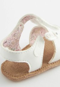 Sandale en faux cuir blanc avec doublure à motifs floraux roses et semelle brune texturée. Présente une sangle Velcro pour un ajustement réglable.