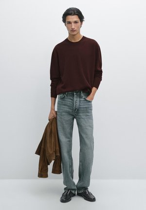 Brunstickad tröja, ljusblå denimjeans, brun mockajacka hålls i ena handen, svarta skor, modell som står framför en enkel bakgrund.