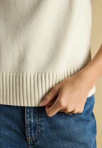 Beiger Strickpullover mit geripptem Saum, der eine glatte Textur zeigt. Hand nahe dem unteren Rand positioniert, kombiniert mit blauen Jeans.