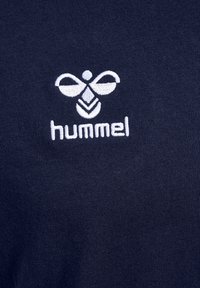 Logo Hummel brodé blanc avec une abeille stylisée au-dessus du texte sur un fond en tissu bleu marine.