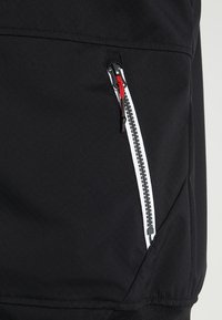 Veste noire avec une poche zippée munie d'un curseur rouge, une fermeture éclair blanche contrastante et un tissu texturé. Coupe ajustée avec une finition lisse.